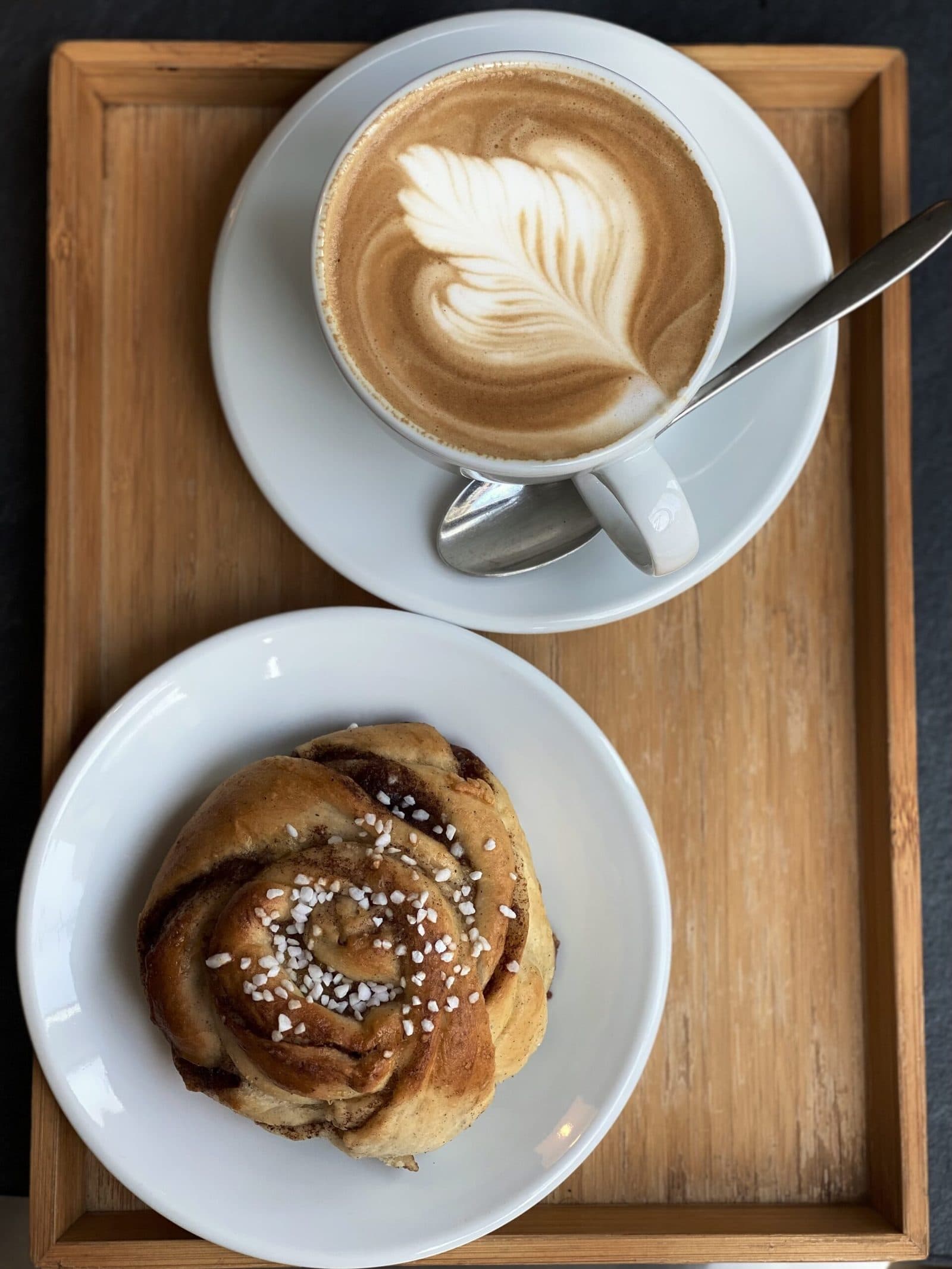 Holztablett mit einer Kanelbulle und einem Cappuccino mit Latte Art in Anns Café